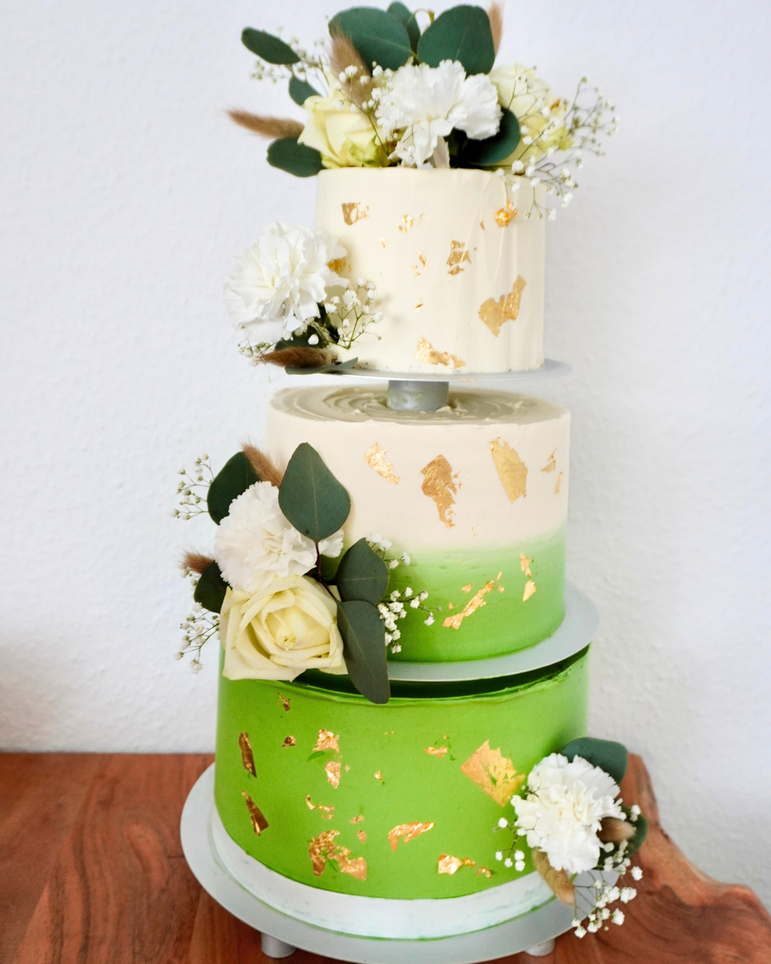 Dreistöckige Torte mit grün-weißer Farbverläufen, verziert mit goldenen Blattgoldakzenten und frischen Blumen wie weißen Nelken, Rosen, Eukalyptus und Schleierkraut