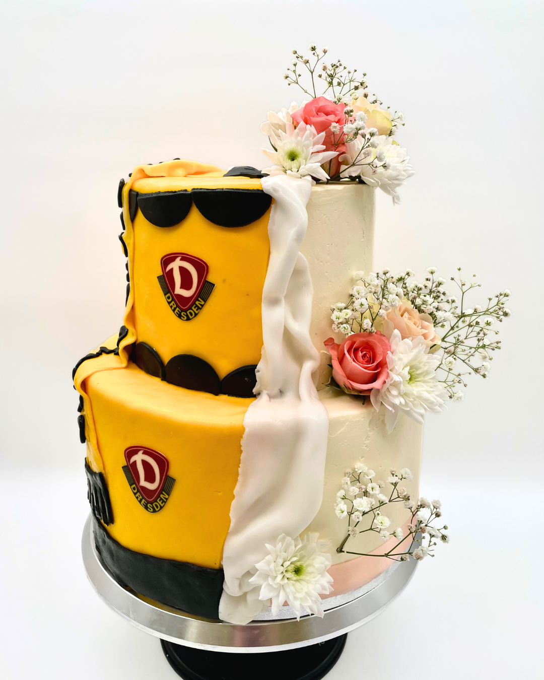 Zweiteilige Torte im Half-and-Half-Design: eine Seite im Dynamo-Dresden-Fußballstil mit gelb-schwarzer Deko und Vereinslogo, die andere Seite in zartem Weiß-Rosa mit romantischen Blumenarrangements aus Rosen, Chrysanthemen und Schleierkraut.