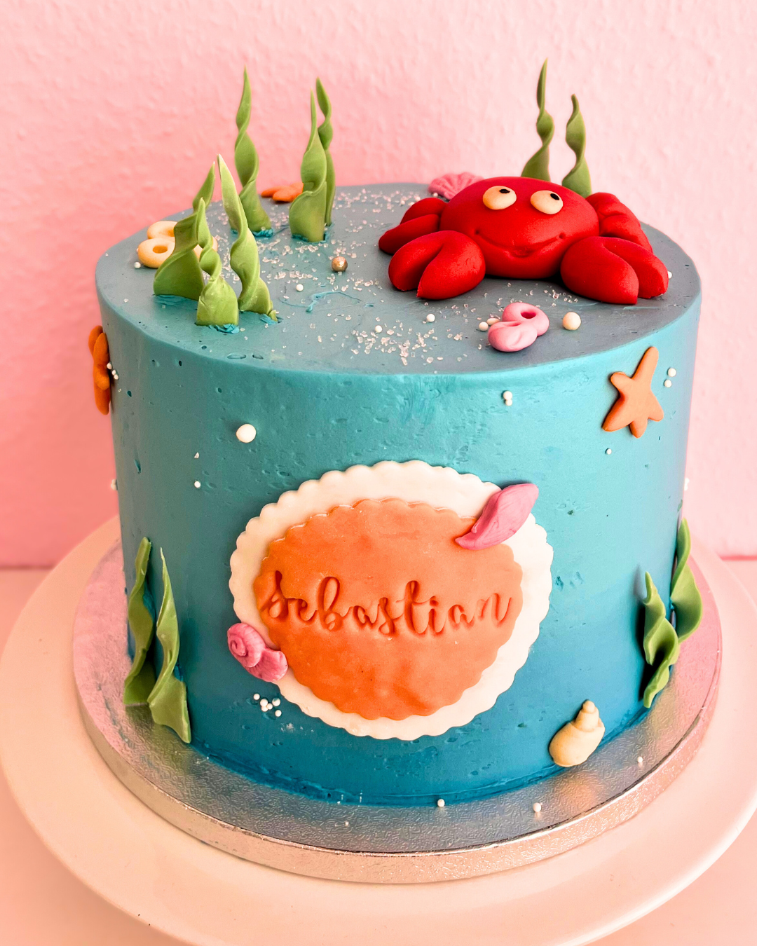 Runde, blau eingestrichene Torte mit Meeresmotiv, dekoriert mit Seegras, Muscheln, Seesternen und einer roten Fondant-Krabbe oben drauf. Auf der Vorderseite steht der Name ‚Sebastian‘ in einer orangefarbenen Plakette