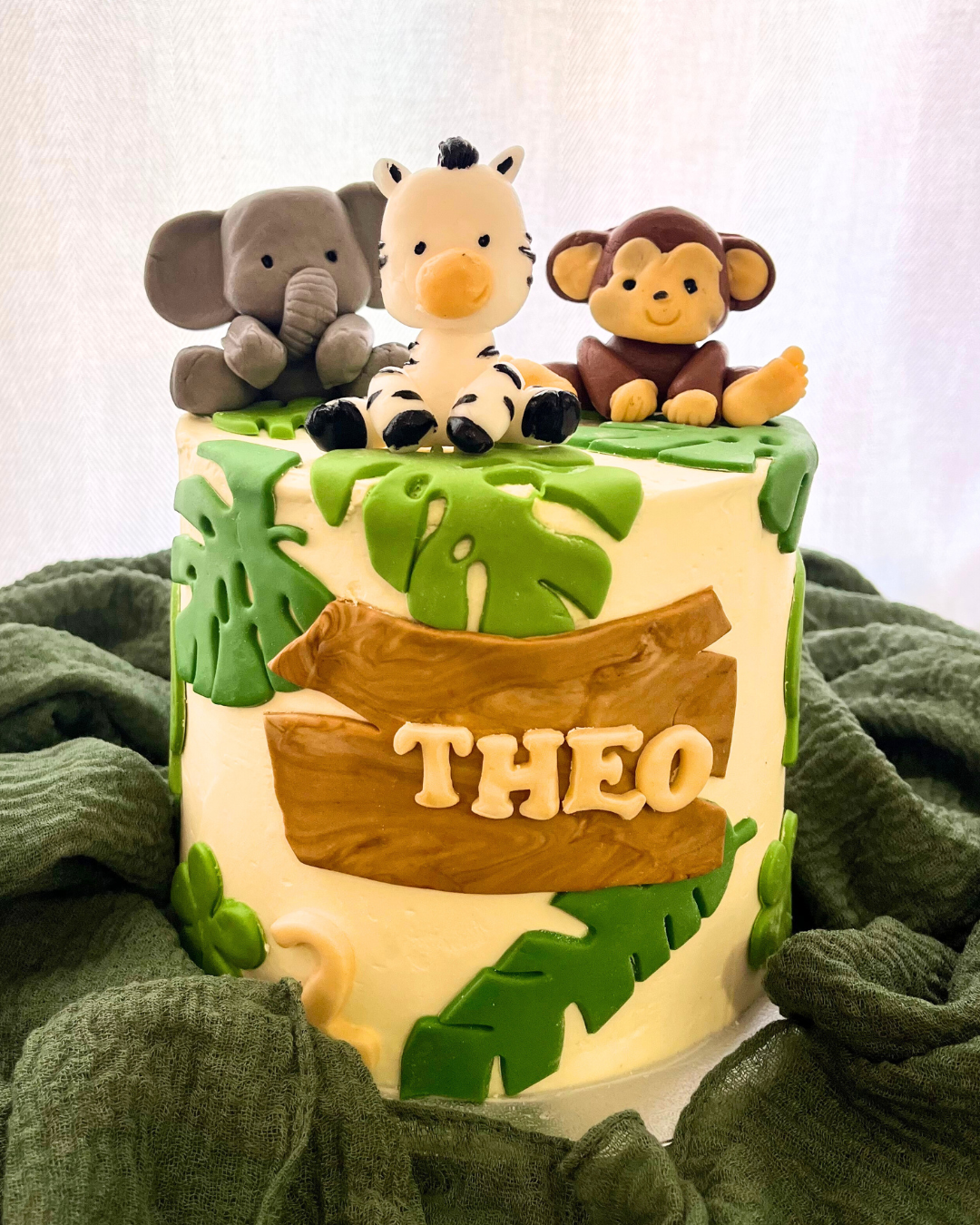 Weiße Torte im Dschungel-Design mit grünen Blättern und einem Holzschild aus Fondant mit dem Namen ‚Theo